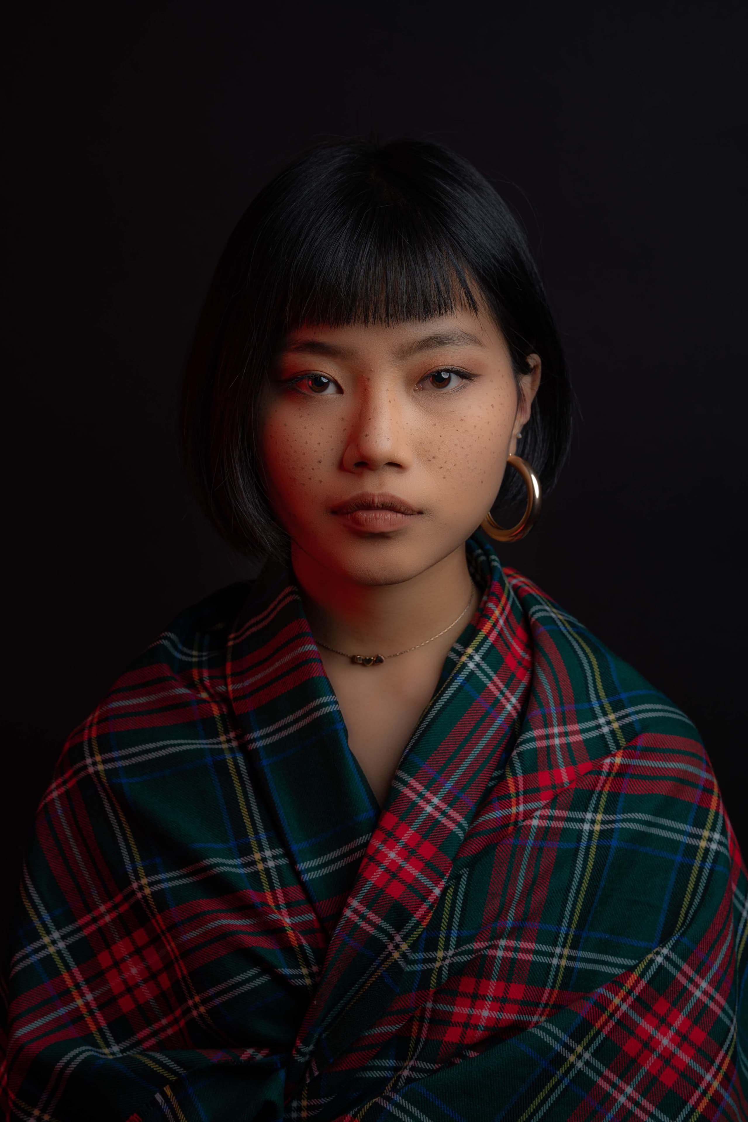 Fashion editorial portrait — model wrapped in tartan against deep shadow, direct gaze