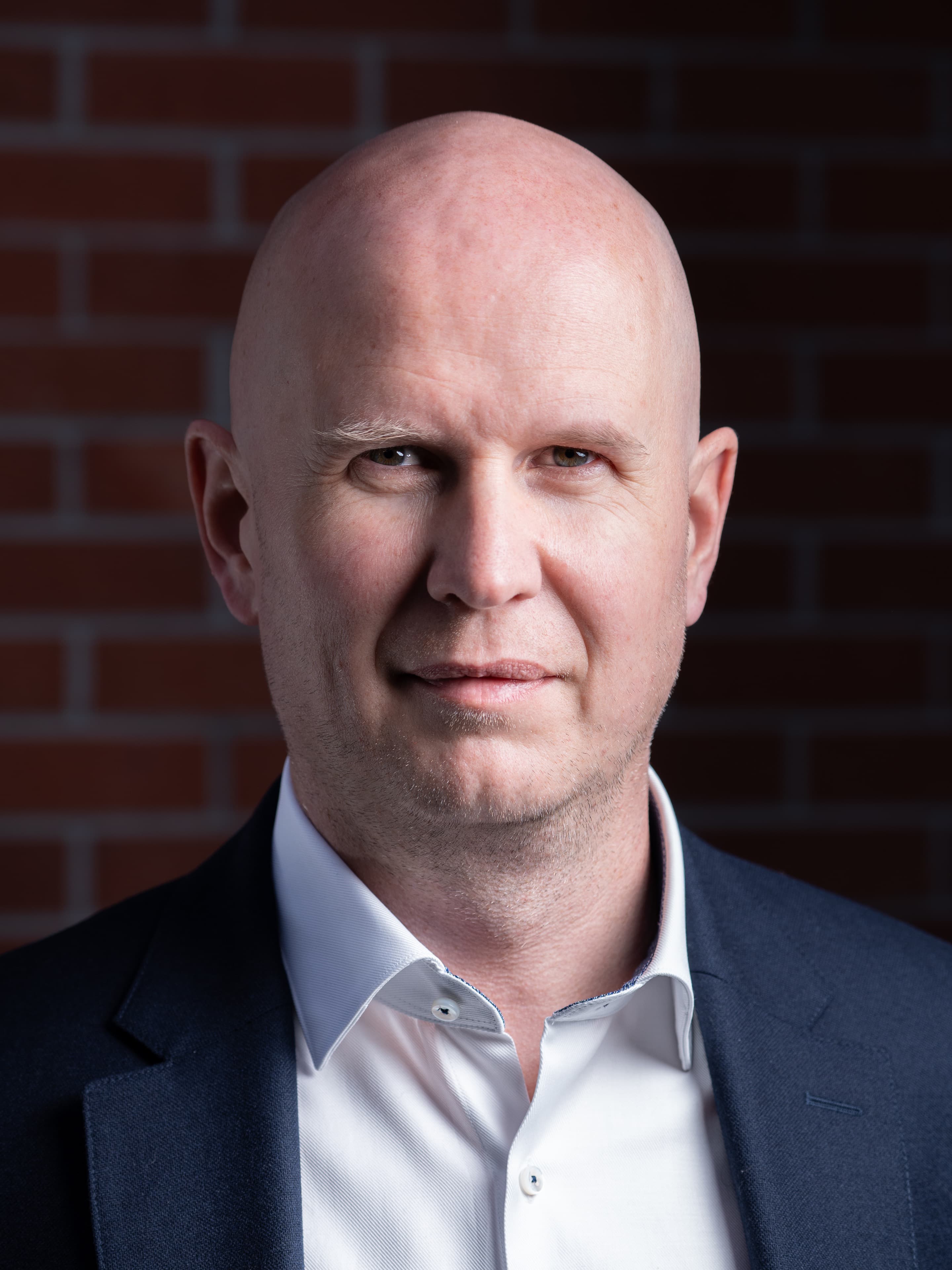 Executive portrait of a bald man in a navy blazer against an exposed brick wall