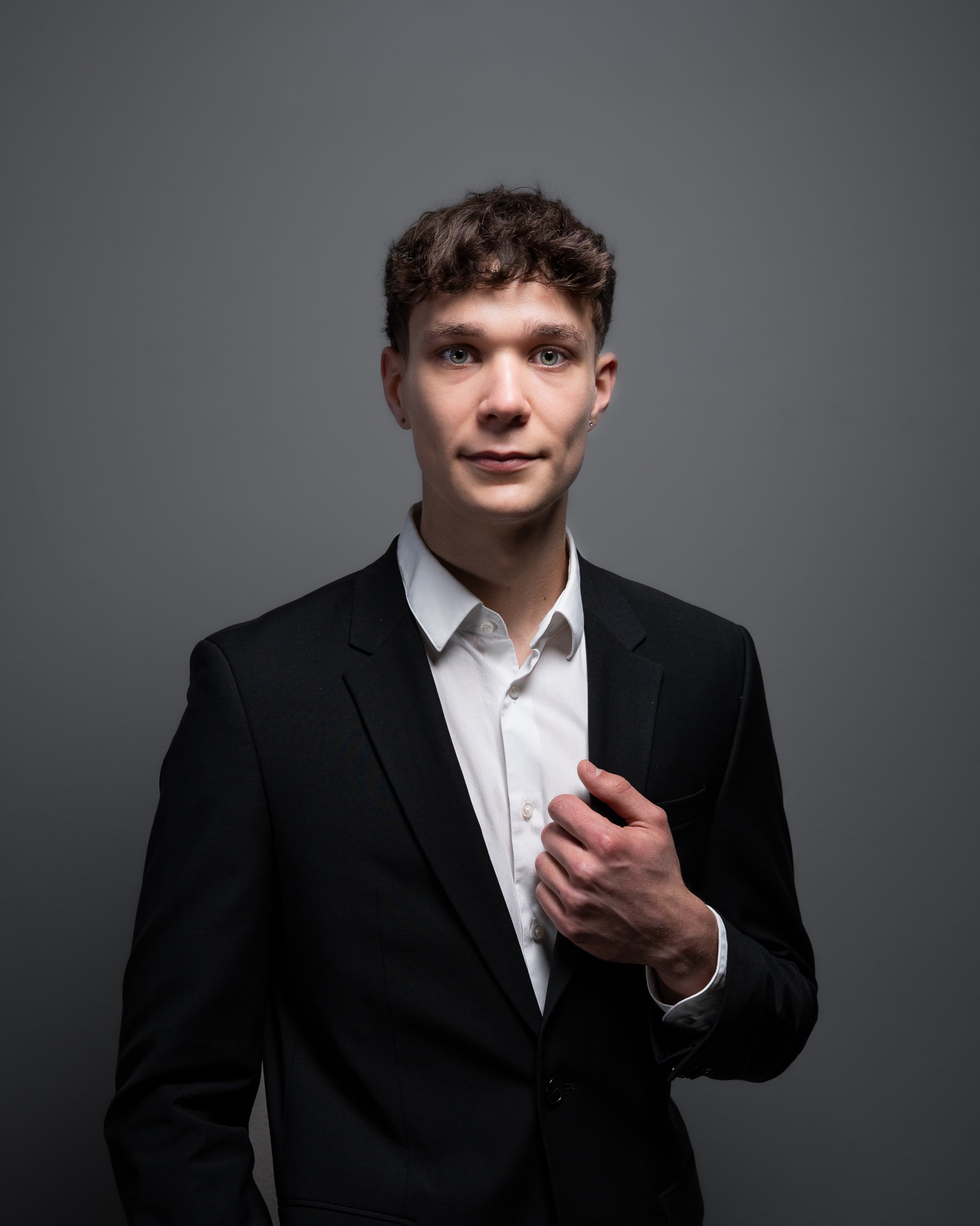 Portrait of a man in a charcoal suit adjusting his lapel on a grey backdrop